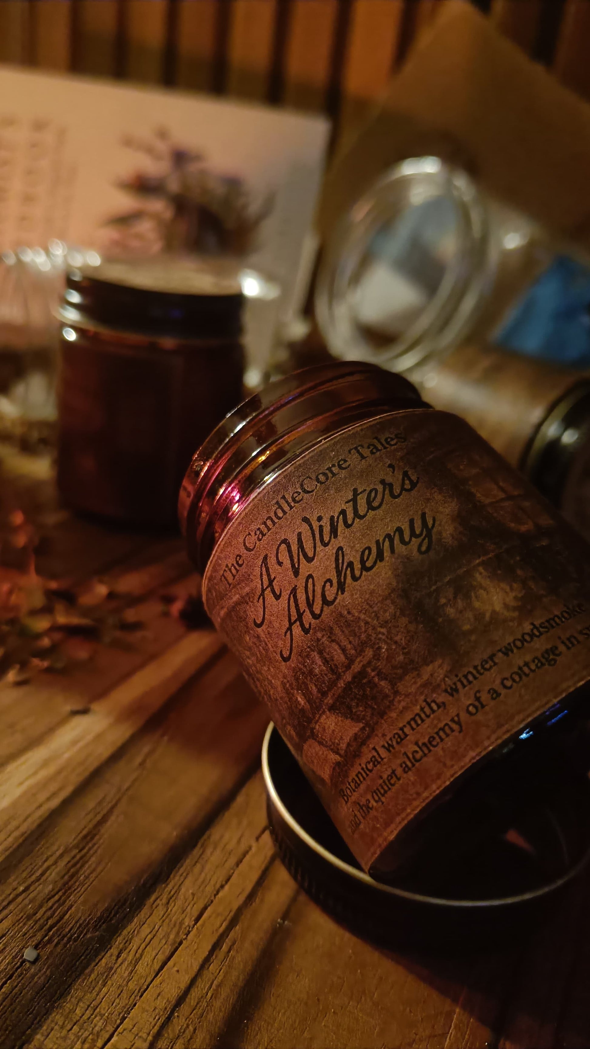 Close-up of A Winter's Alchemy soy candle label illuminated by warm firelight on a rustic wooden surface.