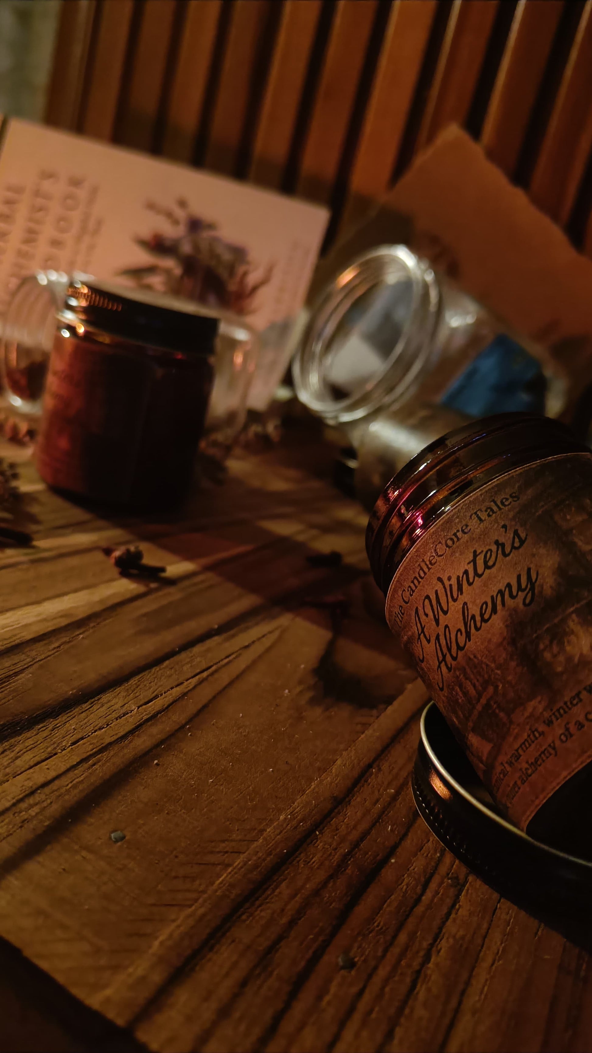 A Winter's Alchemy handmade soy candle resting on a wooden table in lowlight with herbs and books in low light, evoking a quiet mystica mediaeval winter ritual with wam shadows.